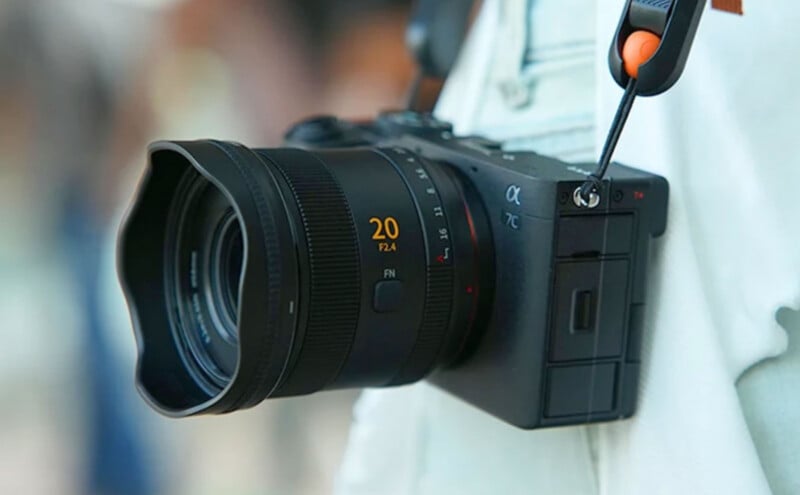 A black mirrorless camera with a 20mm f/2.4 lens hangs from a strap against a person wearing a white shirt.