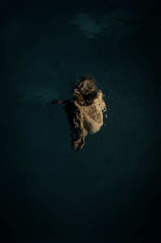 A person with long hair is submerged in dark blue water, with bubbles rising around their head and shoulders.