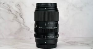 A black Sigma 50-120mm f/2.8 DC OS lens stands vertically against a white marble background.