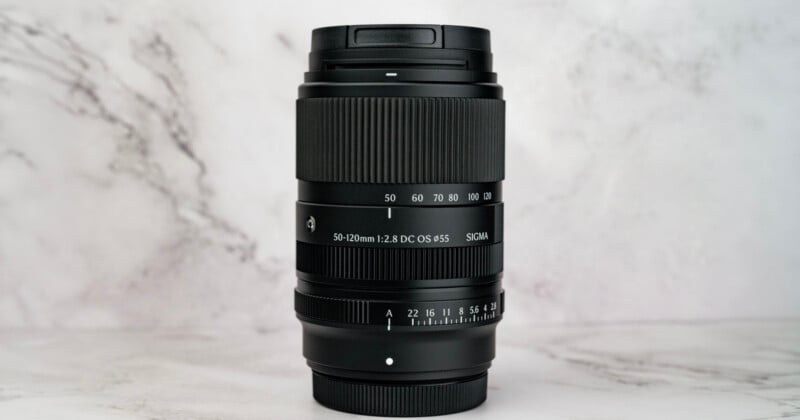 A black Sigma 50-120mm f/2.8 DC OS lens stands vertically against a white marble background.