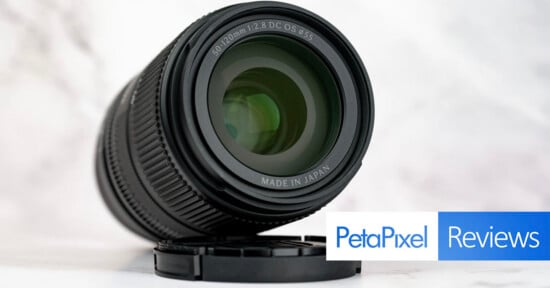 A black camera lens sits on a white surface next to its lens cap, with a PetaPixel Reviews logo in the bottom right corner.