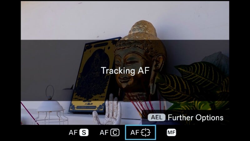 A digital camera screen displays a menu for autofocus settings, with "Tracking AF" highlighted over a view of a golden Buddha statue, a framed print, and a small plant.