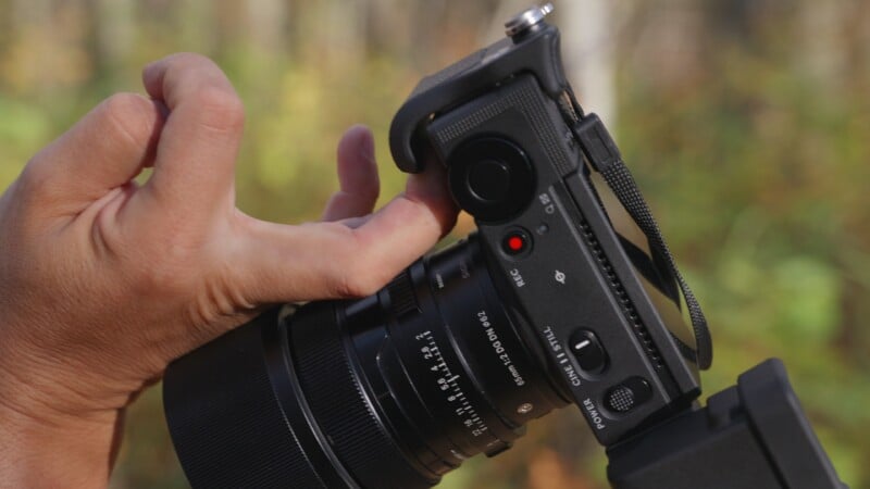 A person's hand holds a black digital camera with a Sigma 65mm lens, showing the top control panel with a red recording button and power switch.