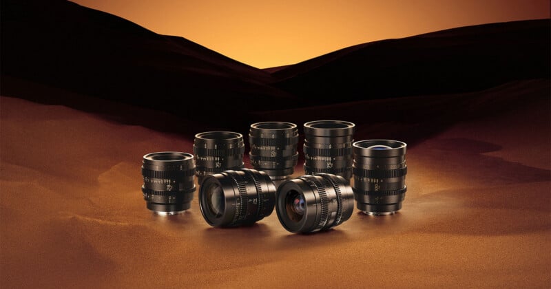Seven black cinema lenses are arranged in a group on a sandy surface against a backdrop of dark, rolling hills under an orange sky.