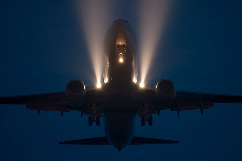 A commercial airplane is captured from directly below during a night landing, with its bright landing lights illuminating the dark sky.