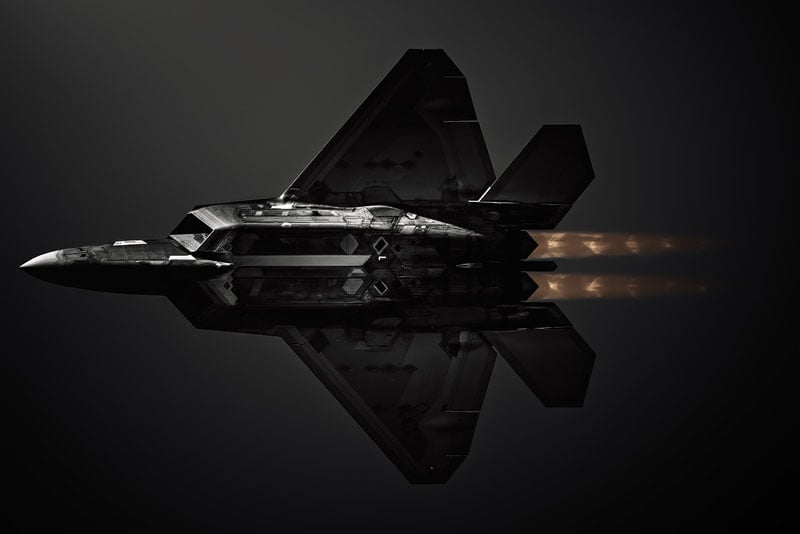 A dark, stealth fighter jet viewed from below against a black background, with visible engine exhaust flames trailing behind.