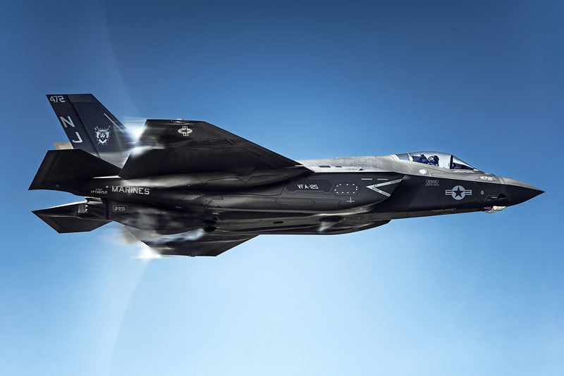 A dark gray F-35 fighter jet with "MARINES" and "VFA-125" markings flies against a clear blue sky.