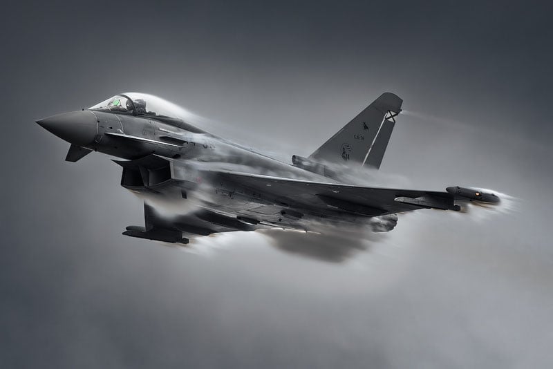 A Eurofighter Typhoon fighter jet flying through humid air, creating visible condensation clouds over its wings and fuselage.