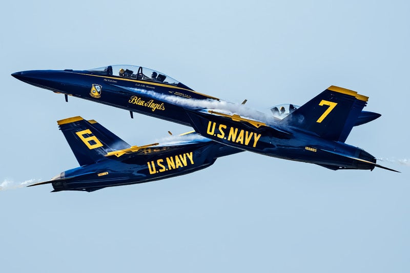 Two U.S. Navy Blue Angels F/A-18 Super Hornets fly in close formation against a clear sky, with the aircraft numbered 7 and 6 visible.