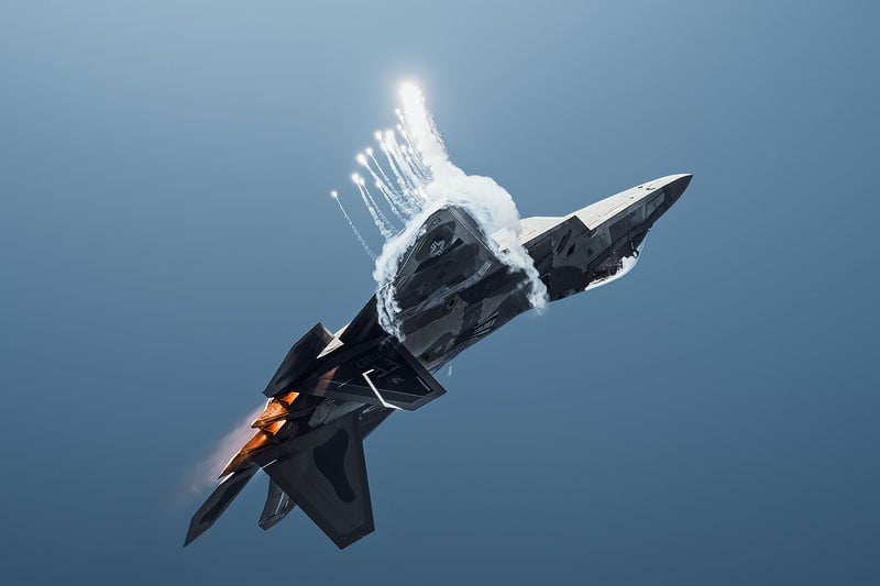 An F-22 Raptor fighter jet performs an aerial maneuver while deploying a series of bright flares against a clear blue sky.