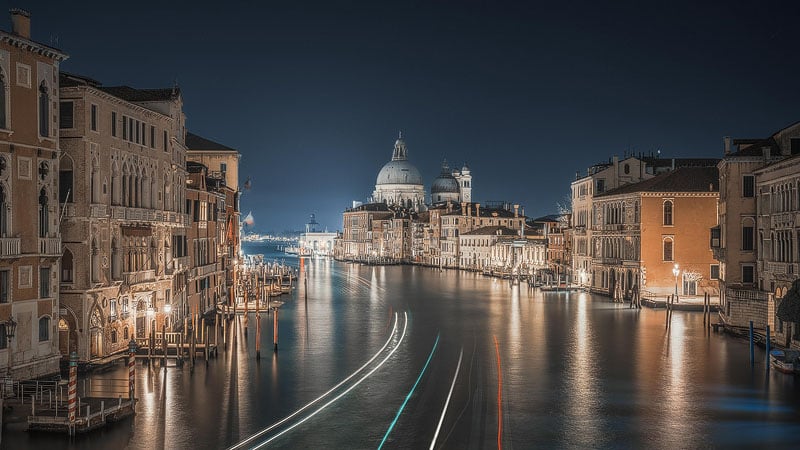 A long-exposure night view of the Grand Canal in Venice, featuring light trails from passing boats and the illuminated Basilica di Santa Maria della Salute in the distance.