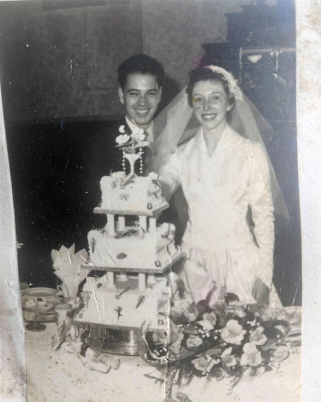 A black-and-white wedding portrait of a smiling bride and groom standing behind a tiered cake.