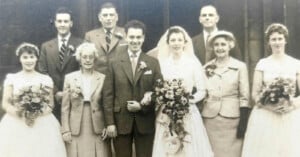 A black-and-white wedding portrait featuring a bride and groom standing in the center, surrounded by four bridesmaids and two older adults.