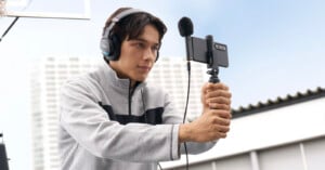 A young man wearing headphones holds a smartphone mounted on a small tripod with an external microphone attached.