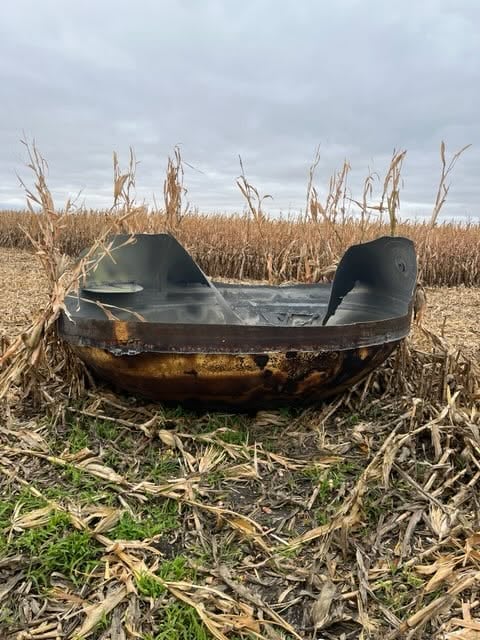 A large, charred, bowl-shaped metal object sits in a harvested cornfield under a cloudy sky.
