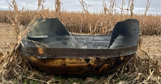 A large, charred, bowl-shaped metal object sits in a field of dry, harvested corn stalks under a cloudy sky.