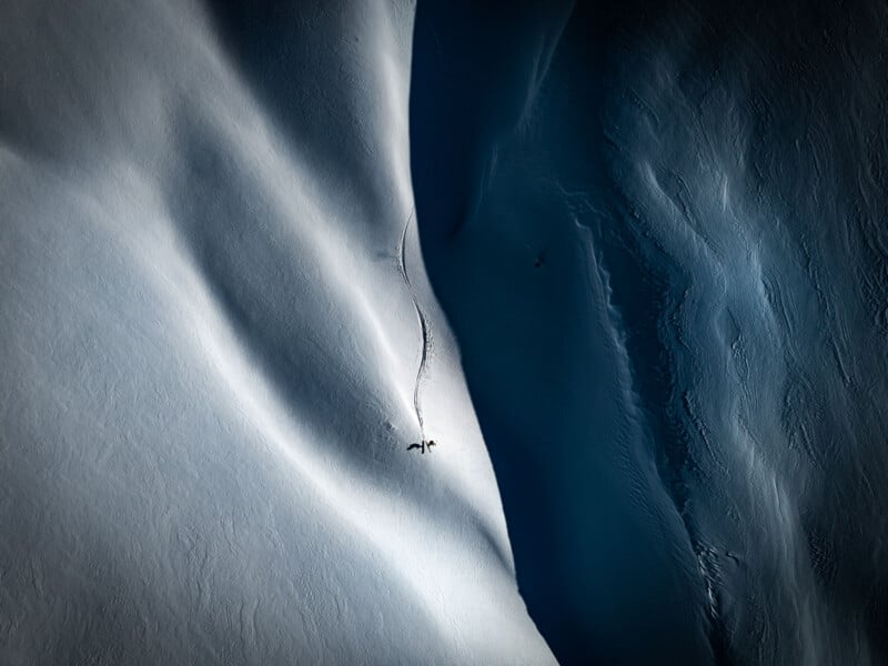 An aerial view shows a lone skier carving a single track down a steep, snow-covered mountain slope next to a deep, dark shadow.