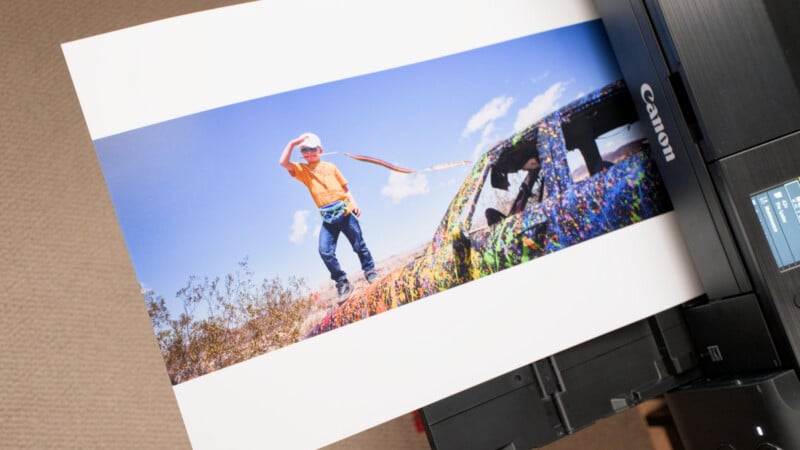 A Canon printer ejects a photograph showing a child in an orange shirt standing on a brightly painted, abandoned car in a desert landscape.