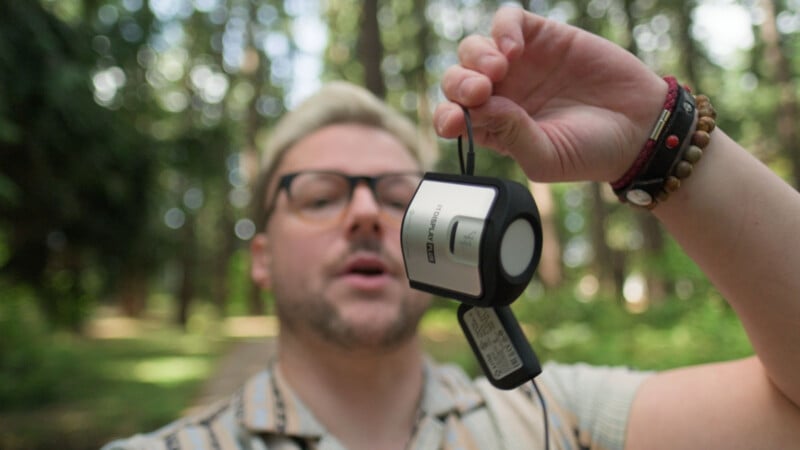 A person with blonde hair and glasses holds up a silver and black X-Rite i1Display Plus color calibration device in a wooded area.