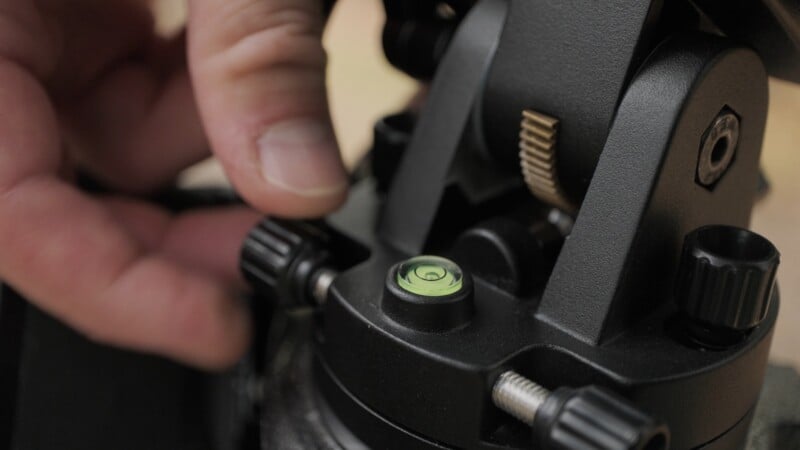 A hand adjusts a black knob on a tripod head next to a small, circular bubble level.