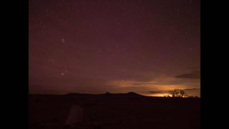 A night sky filled with stars over a dark desert landscape with distant mountains and a low glow on the horizon.