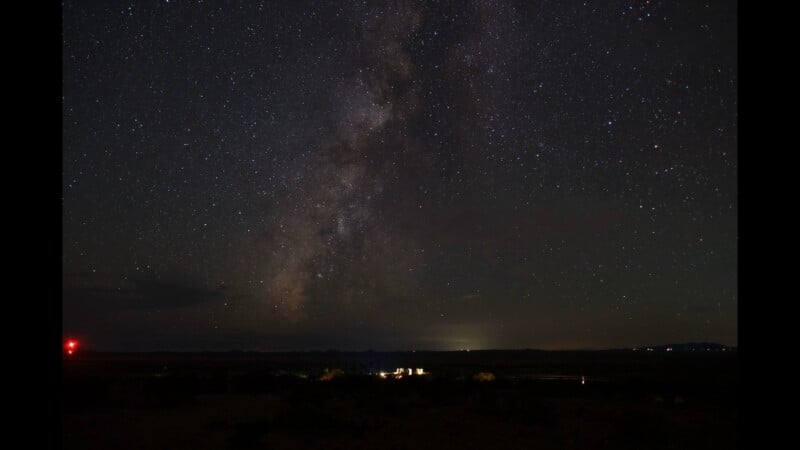 The Milky Way galaxy stretches across a dark night sky above a flat, silhouetted desert landscape with distant lights.