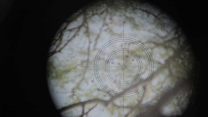 A view through a circular reticle with crosshairs and concentric scales, superimposed over blurry tree branches.