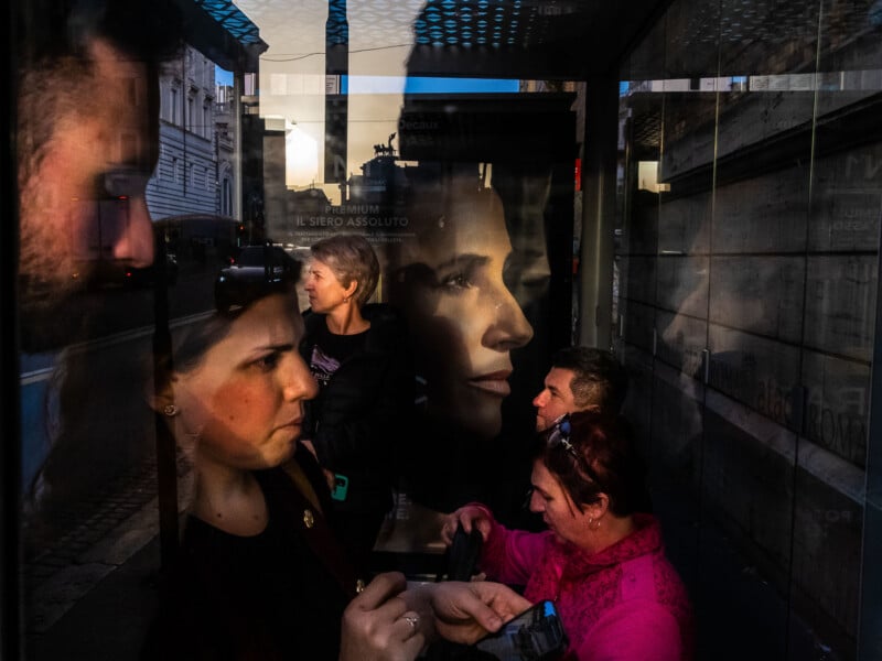 People wait at a glass-walled bus stop, their reflections overlapping with a large advertisement featuring a woman's face.