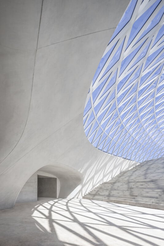 A wide, curved concrete structure features a large skylight with a geometric grid pattern, casting shadows onto a set of stairs and a smooth floor.