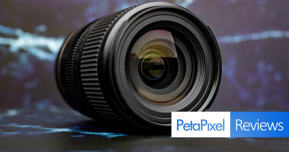 A black camera lens sits on a dark, textured surface against a blurred blue and white background, with a PetaPixel Reviews logo in the bottom right corner.