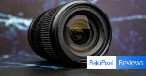 A black camera lens sits on a dark, textured surface against a blurred blue and white background, with a PetaPixel Reviews logo in the bottom right corner.