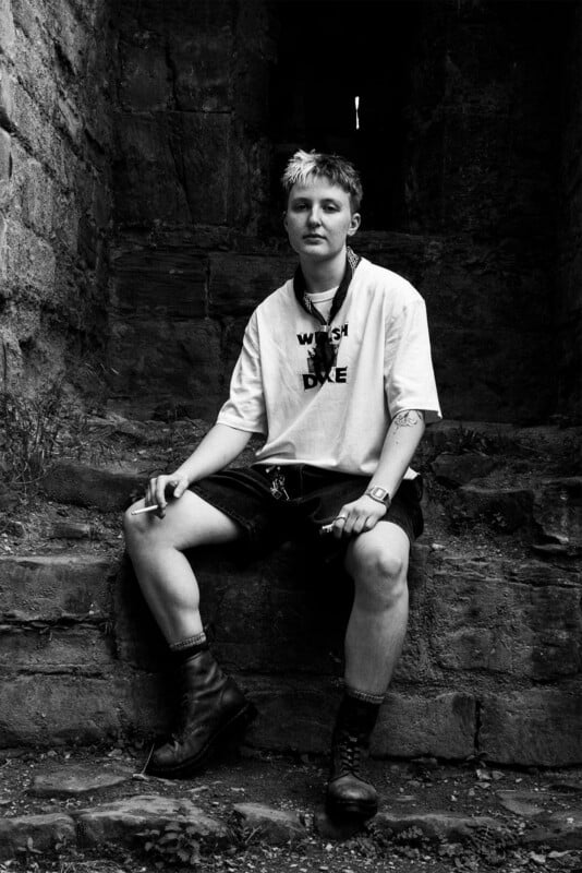 A person with short, two-toned hair sits on stone steps, wearing a white t-shirt, dark shorts, and boots, holding a cigarette.