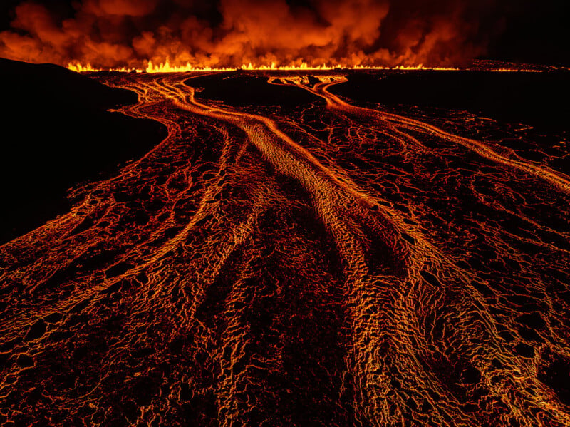 A wide, glowing river of molten lava flows across a dark, volcanic landscape toward a distant line of fire and smoke.