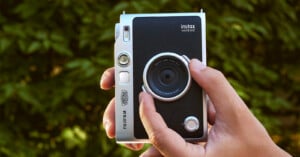 A hand holds a silver and black Fujifilm Instax mini Evo2 camera against a blurred green leafy background.