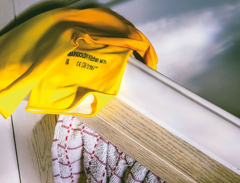A yellow Marigold Kitchen rubber glove rests on a wooden shelf above a white and red checkered kitchen towel.