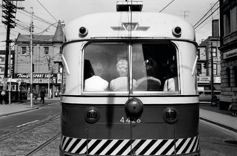 The rear view of a Toronto Transit Commission streetcar, numbered 4426, shows four passengers sitting inside, with city storefronts and street signs visible in the background.