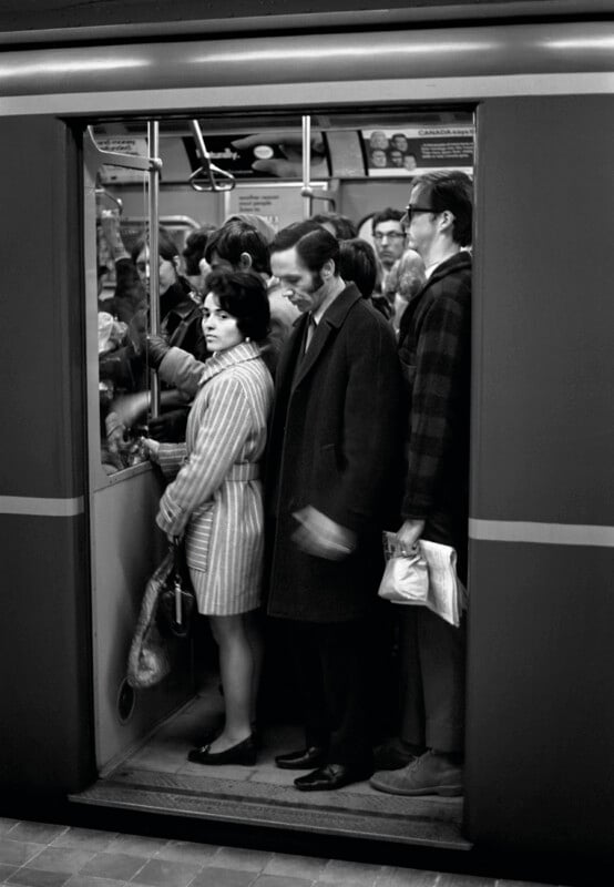 A group of people stand inside a subway car, with a woman in a striped coat looking toward the camera near the open doors.