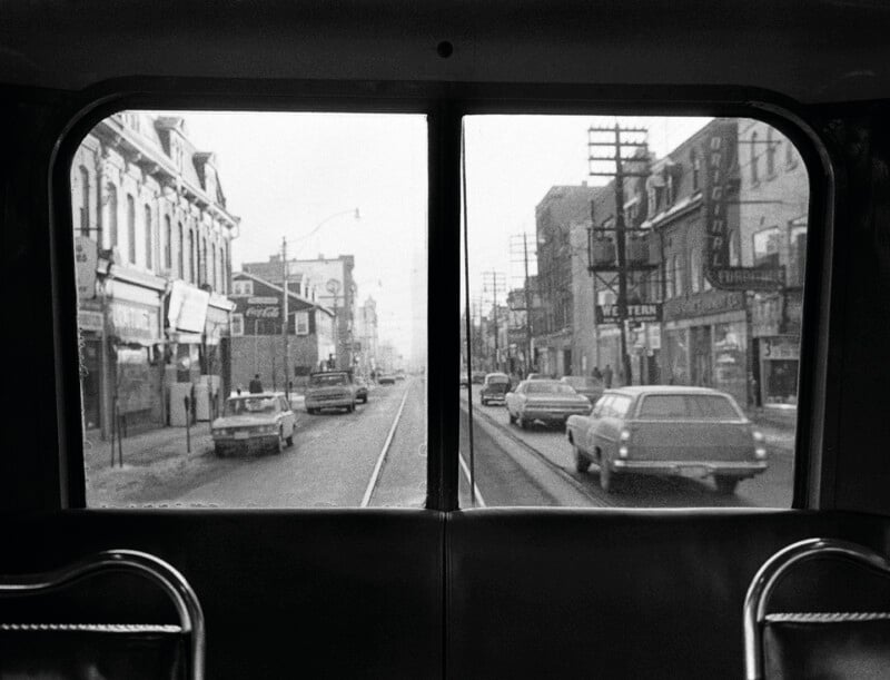 A view from the back seat of a streetcar looking out through two windows at a city street with cars and buildings.