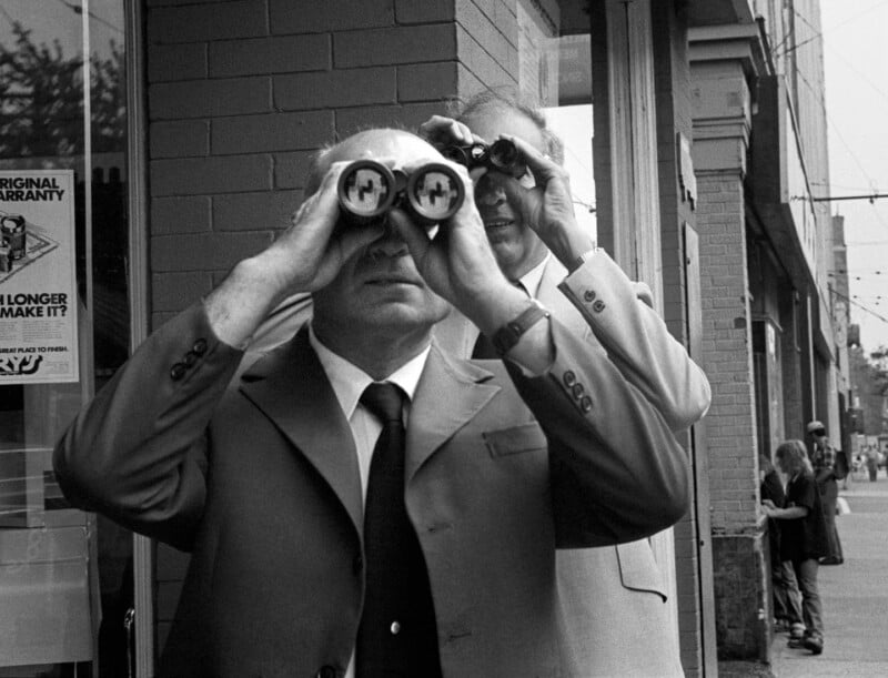 Two men in suits stand on a sidewalk, both looking through binoculars, while a person walks in the background.
