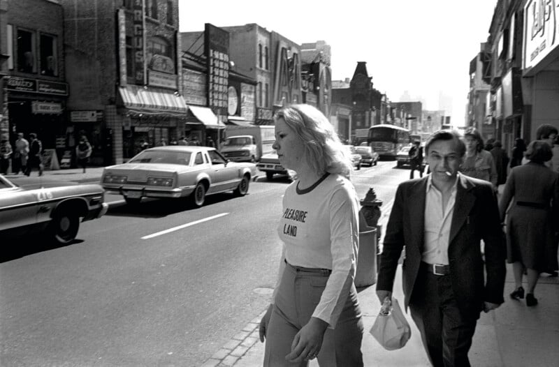 A woman in a "Pleasure Land" shirt and a man with a cigarette walk along a busy city sidewalk past parked cars and storefronts.