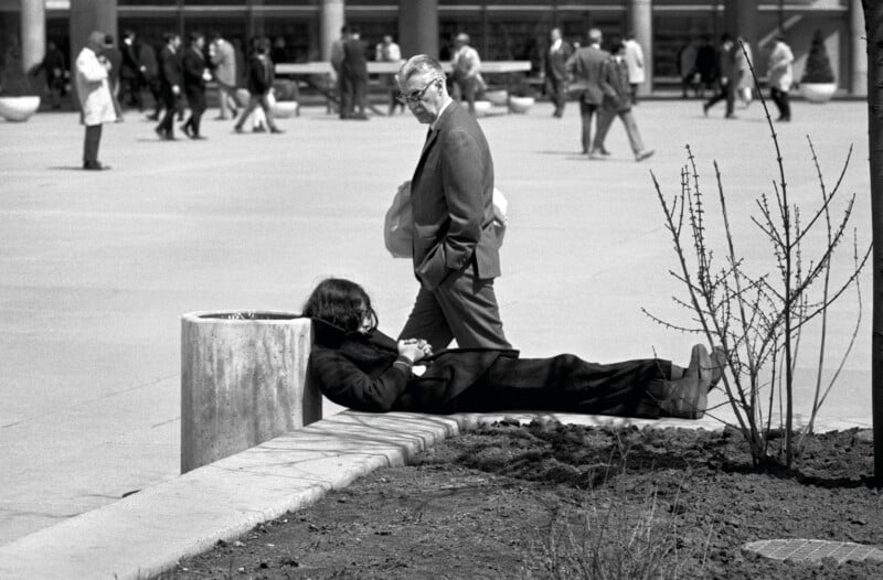 A person with long dark hair lies resting on a concrete ledge next to a trash bin while a man in a suit walks past in the background.