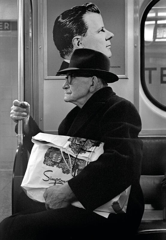An elderly man in a dark coat and fedora sits on a subway train holding a shopping bag, positioned in front of a poster of a man's profile.