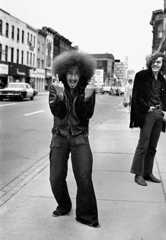 A person with a large afro and a leather jacket stands on a sidewalk, shouting and gesturing with both middle fingers, while another person stands nearby smiling.