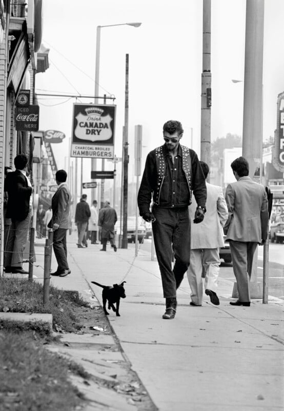 A man in a studded vest and sunglasses walks a small black puppy on a leash down a city sidewalk past several men in suits.