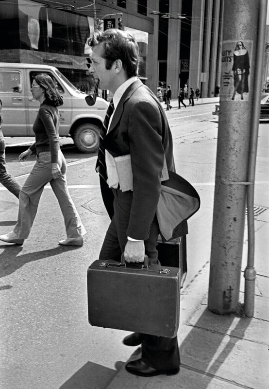 A man in a suit carrying a briefcase and smoking a pipe walks past a woman on a city street.