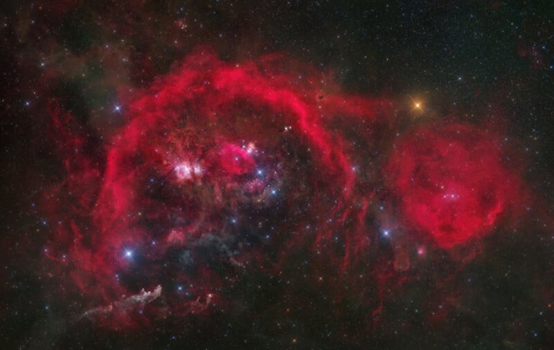 A vast, glowing red nebula fills a dark star-studded sky, featuring complex gaseous filaments and bright clusters of stars.