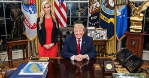 Donald Trump sits at the Resolute Desk in the Oval Office with a woman in a red dress standing behind him, surrounded by military flags and decor.