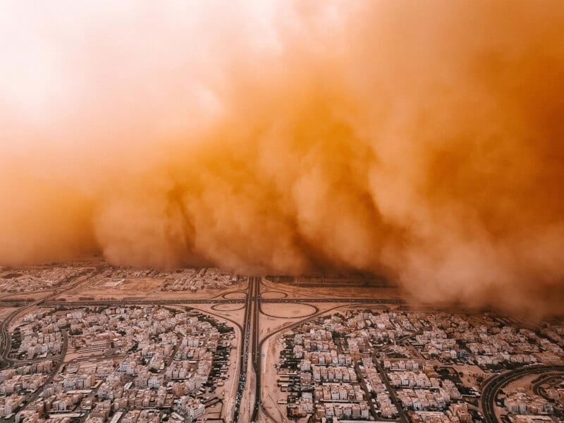 An aerial view shows a massive, dense orange dust storm looming over a suburban landscape and a highway intersection.