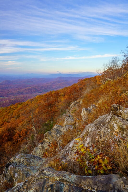A rocky, autumn-colored mountainside overlooks a vast, hazy valley under a blue sky with wispy clouds.