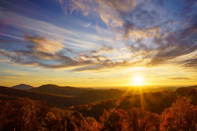 The sun sets over a range of rolling, autumn-colored mountains under a sky filled with wispy, golden clouds.
